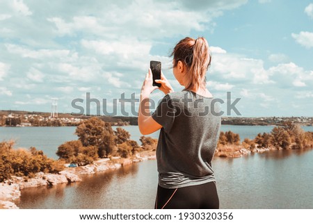 A girl with her hair pulled back stands on the edge of a cliff and takes a picture of the scenic landscape with her phone.