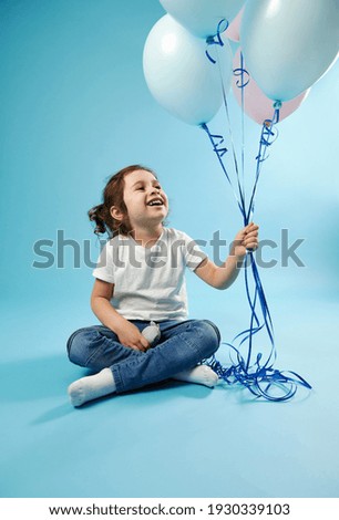 Cute smiling girl sitting on blue background with soft shadow and looking at colored balloons in her hand. Childhood and child protection day concept. Copy space