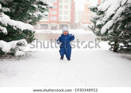 Toddler boy in blue overalls and a cap playing in a snowy spruce forest in winter.