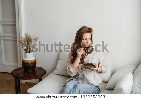 A young blonde woman in casual comfortable clothes sits on a white sofa at home and drinks coffee. scandinavian interior, minimalism