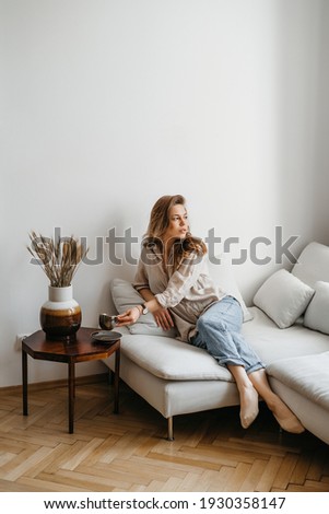 A young blonde woman in casual comfortable clothes sits on a white sofa at home and drinks coffee. scandinavian interior, minimalism Royalty-Free Stock Photo #1930358147