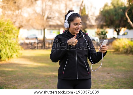 Young brunette sporty girl walking at the park using headphones and smartphone and doing ok sign, thumb up with fingers, excellent sign