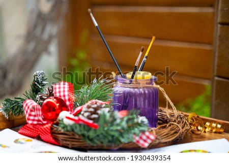 Brushes for painting are in purple glass jar on table decorated with wreath with baubles and ribbons. Artist prepares house for New Year party. Christmas decoration tools. 
