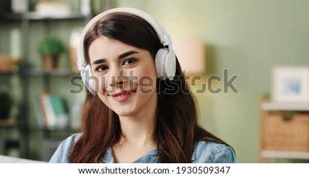 Close up portrait of young beautiful girl sitting at home on couch and enjoying favorite song looking at camera and smiling.