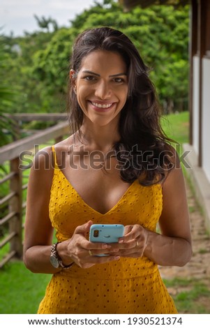 Smiling woman with cellphone in hand and looking at the camera. Countryside landscape in the background.