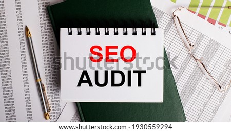 On the desktop are reports, glasses, a pen, a green diary and a white notebook with the words SEO AUDIT. Workplace close-up. Business concept