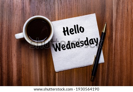 On a wooden table next to a white cup of coffee and a pen is a white paper napkin with the words HELLO WEDNESDAY