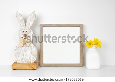 Mock up wood frame with Easter decor on a white shelf. Rustic wood bunny and vase of spring flowers. Square frame against a white wall. Copy space.