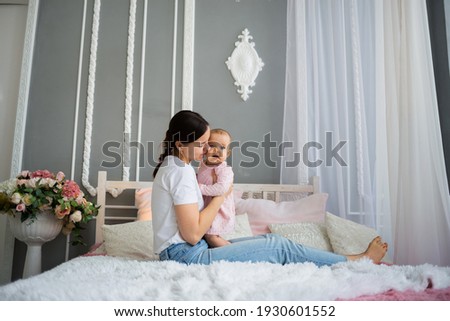 brunette mom in a white T-shirt sits on the bed and holds her baby daughter on the bed