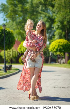 A charming girl in a light summer sundress walks in a green park with her little daughter, holding her in her arms. Enjoys warm sunny summer days.