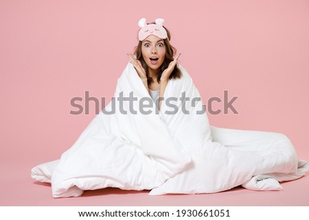 Shocked woman in pajamas home wear sleep mask sit wrapping in blanket keeping mouth open spreading hands rest at home isolated on pink background studio portrait. Relax good mood lifestyle concept