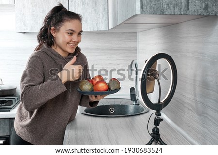 Beautiful Caucasian female blogger shows fruits and clean diet food. In front of the camera to record a video blog, the video is broadcast live at home