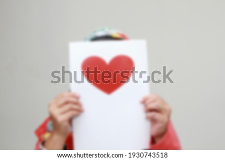 A completely blurry, unfocused portrait of a woman who hides her face behind white paper with a red heart printed on it.