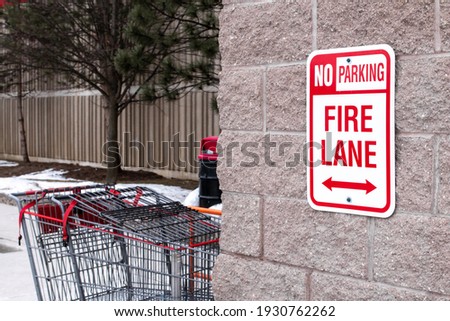 A 'No Parking,' Fire Lane red vertical sign with double headed arrows bolted to a beige brick wall outside a super market in London, Ontario, Canada. Shopping carts and a trash can in the back.