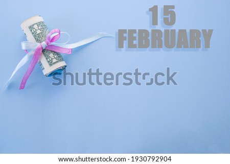 calendar date on blue background with rolled up dollar bills pinned by blue and pink ribbon with copy space. February 15 is the fifteenth day of the month.