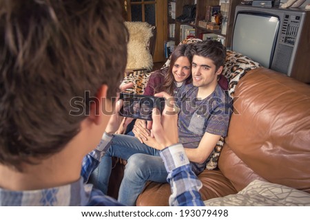 View of young friend taking photos with a smartphone to hipster teenage couple sitting in the sofa