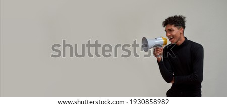 Joyful young guy with piercing dressed in black looking away and screaming in megaphone, making sale announcement, posing isolated over gray background. Advertisement concept. Copy space