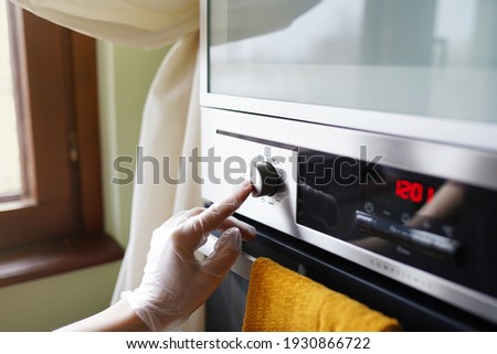 the left hand in a glove presses the button to turn on the oven in the kitchen, a yellow towel hangs next to it, the entire oven panel is clean