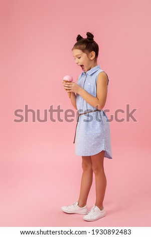 cute smiling little girl eating vanilla ice cream in a waffle cone on pink background. full length