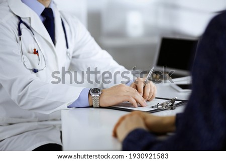 Unknown young woman patient discuss the results of her medical tests with a doctor, while sitting at the desk in a hospital office. Physician using clipboard for filling up medication history records Royalty-Free Stock Photo #1930921583