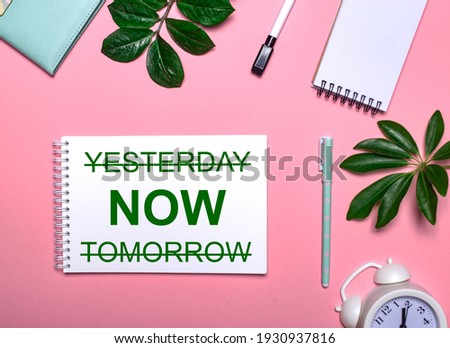 YESTERDAY NOW TOMORROW is written in green on a white notepad on a pink background surrounded by notepads, pens, white alarm clock and green leaves. Educational concept
