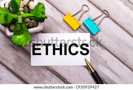 On a wooden background, a white card with the text ETHICS, yellow and green paper clips and a plant in a pot. View from above