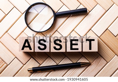 Wooden cubes with the word ASSET stand on a wooden background between a magnifying glass and a pen