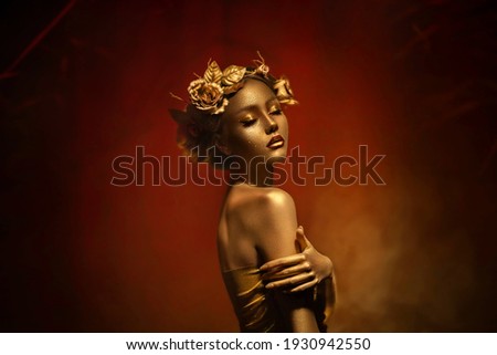 Fantasy portrait of woman with golden skin. Girl goddess in wreath, gold roses, accessories. Beautiful face, steel glitter makeup. Artistic photo dark red background. Elf fairy princess. Fashion model