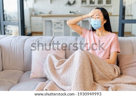 An ill Asian woman wearing face mask sits alone at home covered with a blanket, has fever and feels headache, got a viral infection or flu, isolated at home Royalty-Free Stock Photo #1930949993