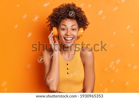Joyful dark skinned curly girl enjoys listening music via wireless headphones smiles gently enjoys spare time dressed in casual top isolated over orange background soap bubbles flying around