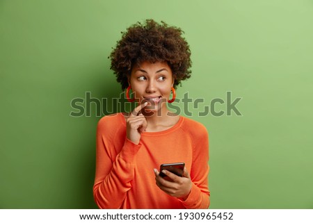 Good looking dark skinned young woman with curly hair holds modern smartphone uses new application dressed in casual orange sweater isolated over green background thinks about something pleasant Royalty-Free Stock Photo #1930965452
