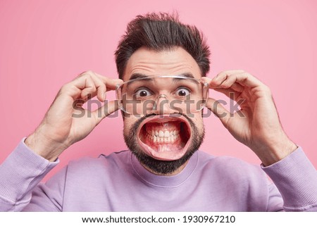 Startled handsome man stares surprisingly through transparent spectacles clenches teeth has widely opened mouth hears great news stands alarmed being super shocked stands indoor. Emotions concept Royalty-Free Stock Photo #1930967210