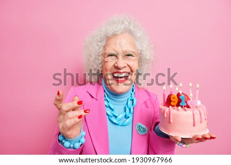 Life only starts when get older. Amused fashionable senior lady celebrates birthday holds cake with burning candles wears makeup isolated over pink background. Positive granny celebrates 91st bday