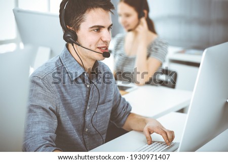Casual dressed young man using headset and computer while talking with customers online. Group of operators at work. Call center