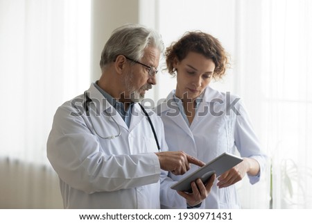 Focused mature male doctor and female nurse look at tablet screen discuss anamnesis together. Concentrated diverse medical professionals use pad device, engaged in team thinking in hospital. Royalty-Free Stock Photo #1930971431
