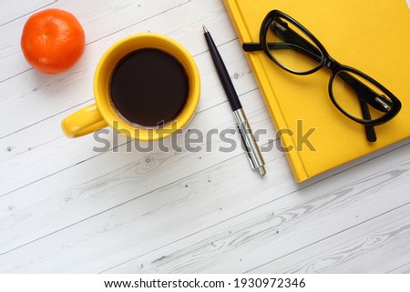 A yellow notebook, a yellow coffee cup, tangerine, glasses, and a ballpoint pen on a wooden background. Close up, top view, diagonal composition. Office picture.