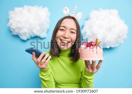 Happy beautiful woman holds birthday cake and smartphone device smiles toothy glad to accept congratulations on her 26th bday poses against blue background with white clouds above. Celebration concept
