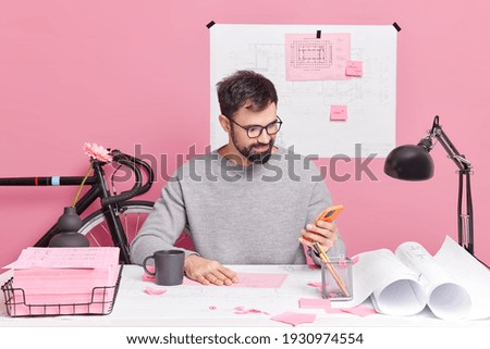 Photo of student engineer does homework makes drawings checks email box via smartphone wears spectacles and jumper poses in coworking space prepares architectural project sits at office desk