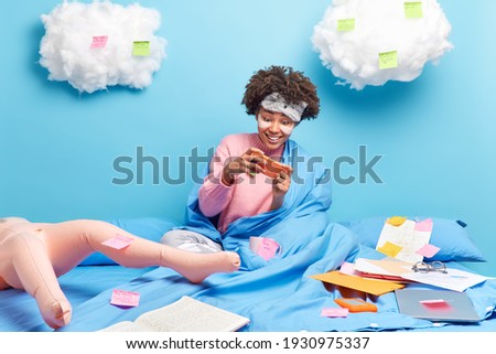 Happy addicted teenage girl plays video games on modern mobile phone has new level rests after exam preparation stays in bed works with papers isolated over blue background with clouds above