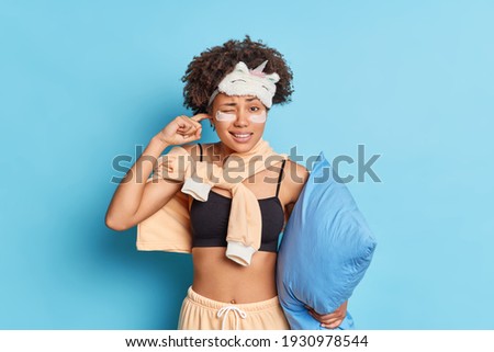 Photo of dark skinned woman plugs ears cannot fall asleep because of noise wears nightwear sleeping mask applies patches under eyes holds pillow isolated over blue background. Sleep interruption