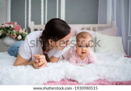 beautiful brunette mom in a white T-shirt lies on the bed with her baby daughter in a pink dress