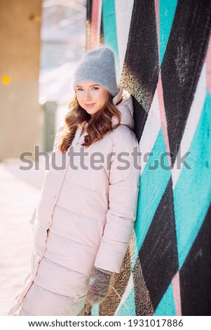 A girl in the winter on the street in the city in a delicate pink jacket smiles while standing next to a painted wall. image with selective focus