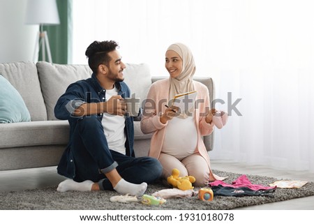 Happy pregnant muslim couple getting ready for childbirth, making checklist of necessities while sitting together on floor in living room with baby clothes around, islamic spouses relaxing at home