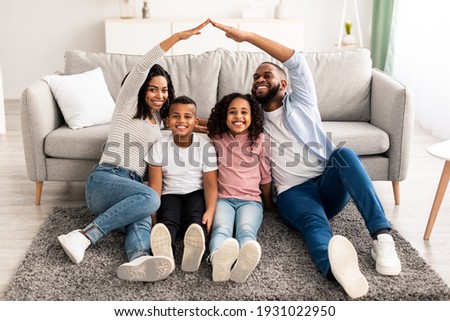 Family Care, Protection And Insurance Concept. Portrait of smiling African American parents making symbolic roof of hands above their happy children, sitting on the floor carpet in living room at home Royalty-Free Stock Photo #1931022950