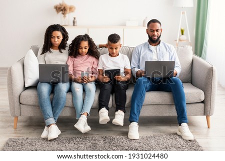 Gadgets Addiction. Portrait of African American family of four people holding and using different electronic devices while sitting on the sofa in living room at home, parents and their little children