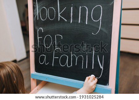 A little girl, a child from an orphanage in search of a family, writes a dream on a wooden black board, easel, holding white chalk in her hand. Photo, copy space, message.