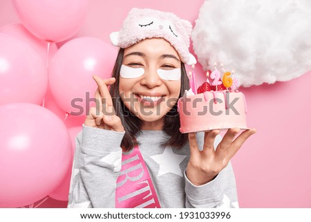Positive Asian woman makes wish before blowing candles on birthday cake crosses fingers smiles gladfully dressed in pajama sleepmask applies beauty pads celebrates 26th bday at home balloons behind