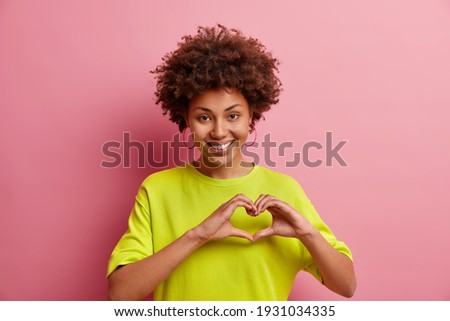 Cute smiling Afro American woman makes I love you gesture confesses in love expresses sympathy shows heart sign dressed in casual clothes isolated over pink background. Body language concept