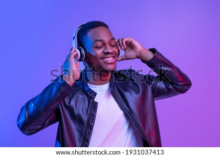 Music Lover. Handsome Black Guy In Wireless Headphones And Leather Jacket Listening His Favorite Songs, Relaxed African American Man Standing With Closed Eyes Under Neon Light Over Purple Background Royalty-Free Stock Photo #1931037413