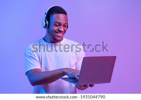 Online Gaming. Portraif Of Happy Black Man In Wireless Headphones Using Laptop Computer, Playing Games In Internet While Standing In Neon Lighting Over Purple Studio Background, Copy Space